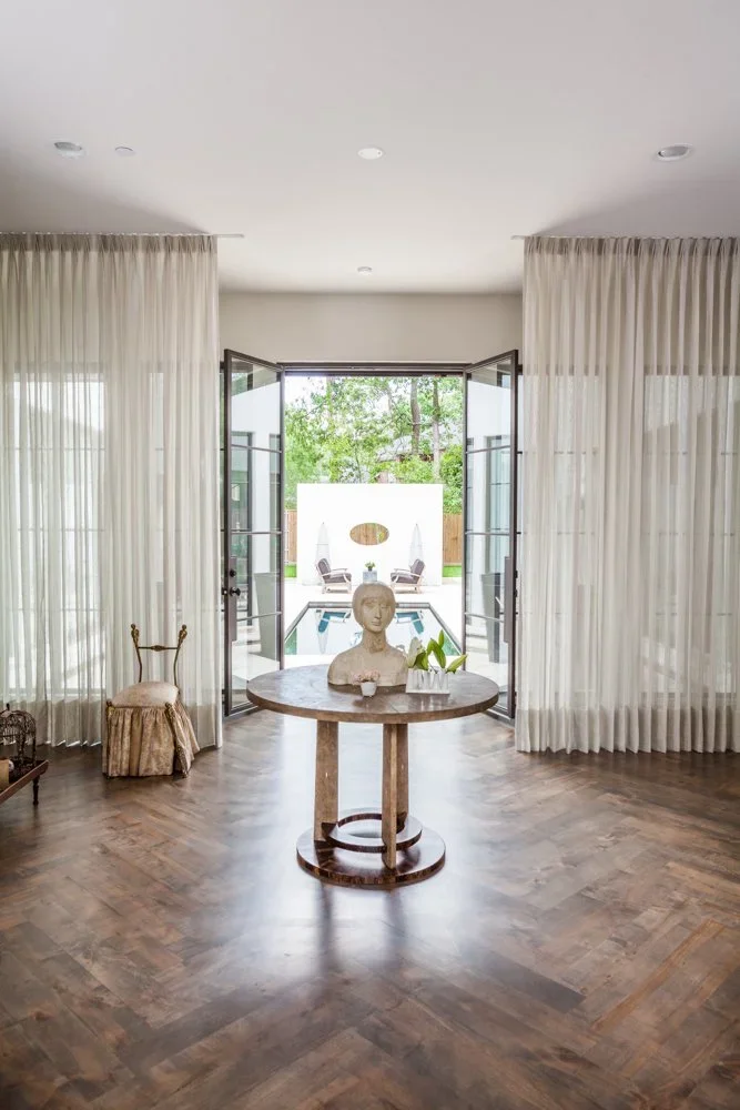 Interior view of a room with an open door leading to an outdoor area with trees and a pool; the room has dark wood flooring, sheer white curtains, a round wooden table with decorative items including a bust sculpture and a plant, and a small upholstered stool.