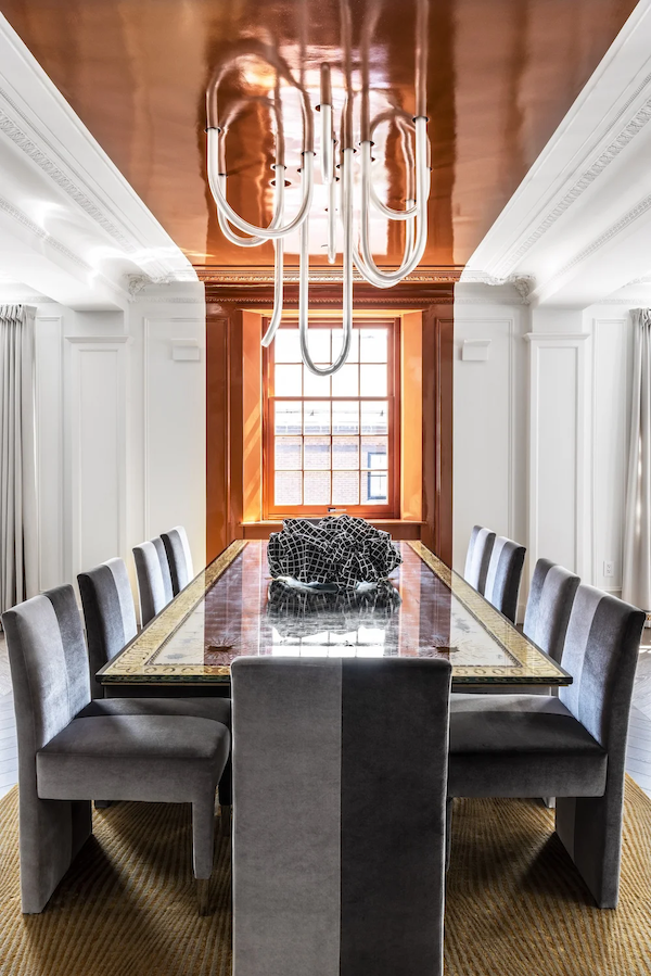 Elegant dining room with a large wooden table, eight gray upholstered chairs, a decorative black and white fabric on the table, white paneled walls, a window with natural light, and a modern, abstract chandelier hanging from a polished wooden ceiling.