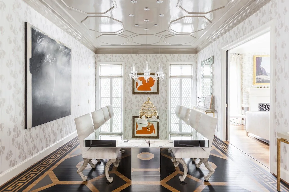 Elegant dining room with a reflective black glass table, eight white upholstered chairs, patterned wallpaper, and a chandelier above. Artworks are on the walls, and a birdcage centerpiece is on the table. Bright windows let in natural light, and a doorway leads to another room.