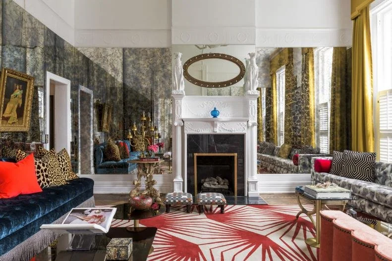 Elegant living room with a white ornate fireplace, large mirror, patterned rug, and mix of modern and classic furniture. The walls are decorated with art and mirrors. A mix of bold colors like red, gold, and blue accents the space.