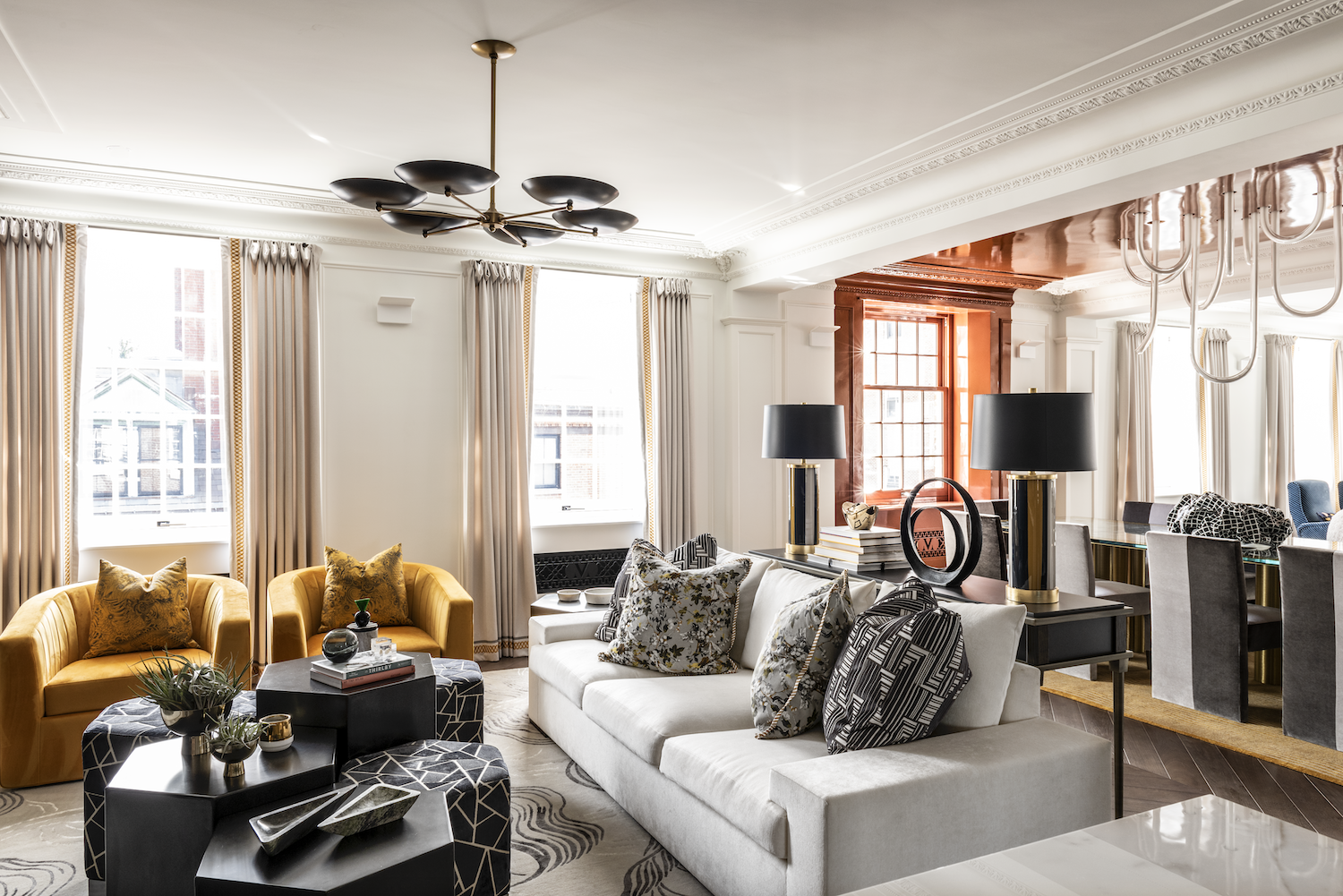 Modern, elegant living room with white walls, large windows with cream curtains, yellow armchairs, a white sofa with decorative pillows, black coffee tables with plants and books, black lamps, and a dining area in the background with gray chairs and large windows.