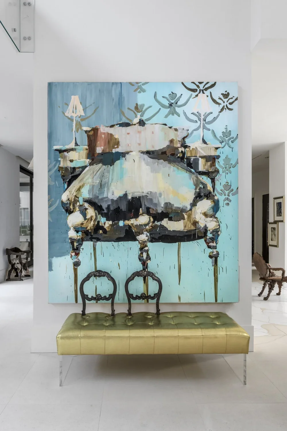 Interior of a modern room with a large abstract painting of a vintage sewing machine, a gold bench with two ornate black chairs in front, and decorative lamps on the painting.